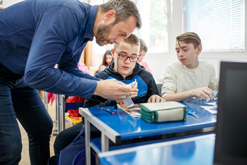 A teacher assists two students who appear to be male with an assignment. The teacher is bent over their desk working intently. One student appears to have a disability.