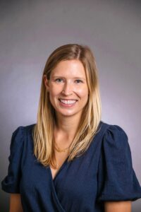 A woman in a blue shirt sits in front of a gray backdrop and smiles