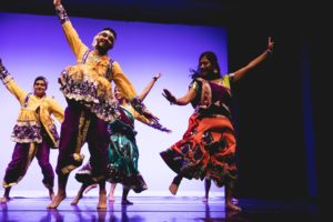 Students wearing brightly-colored costumes perform a dance onstage in front of a purple background 