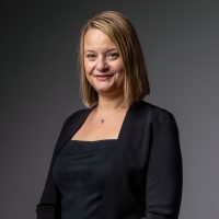 A woman in a black dress and jacket standing against a gray background