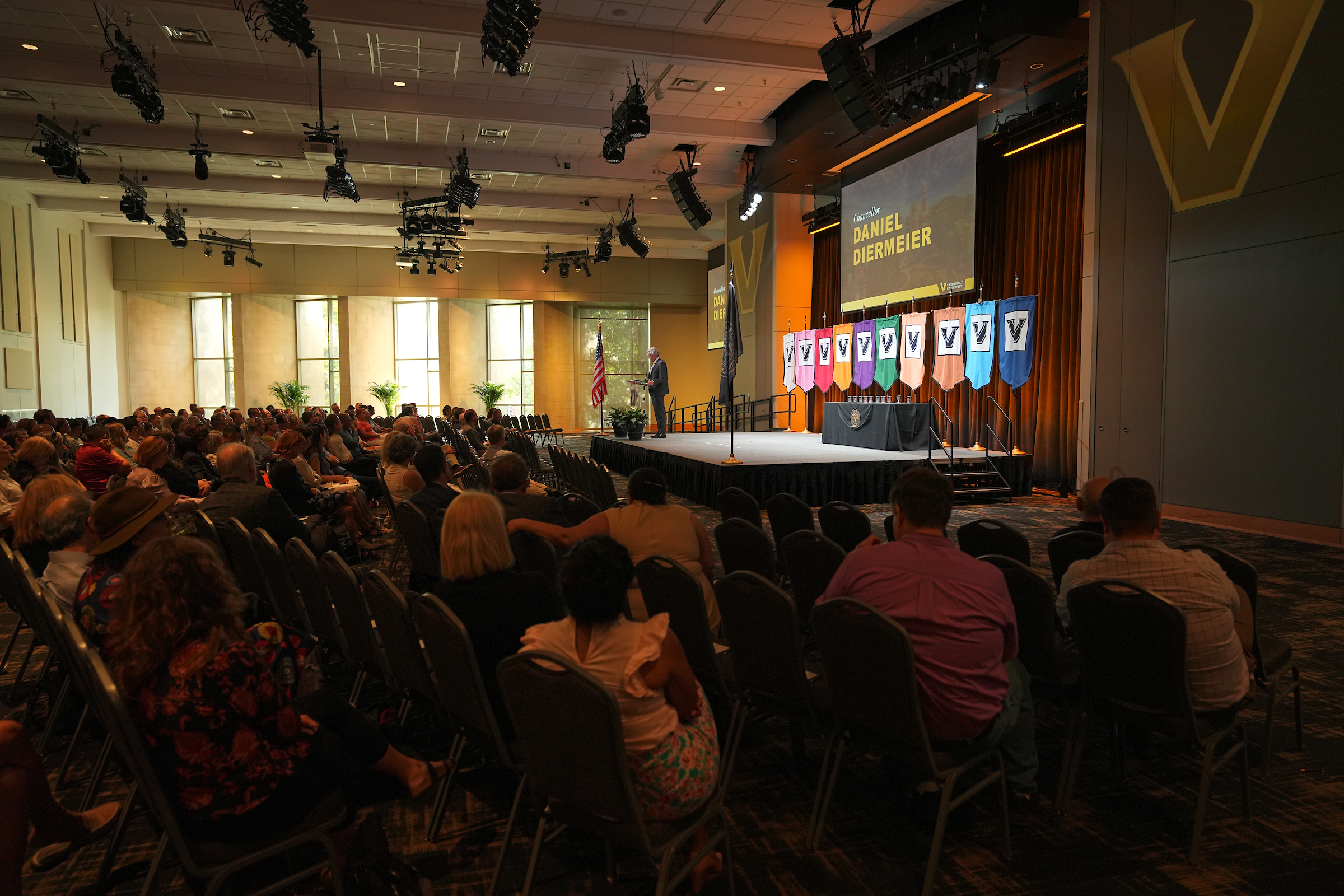 An audience seated in a darkened conference room watching a slideshow presentation and speaker on stage.
