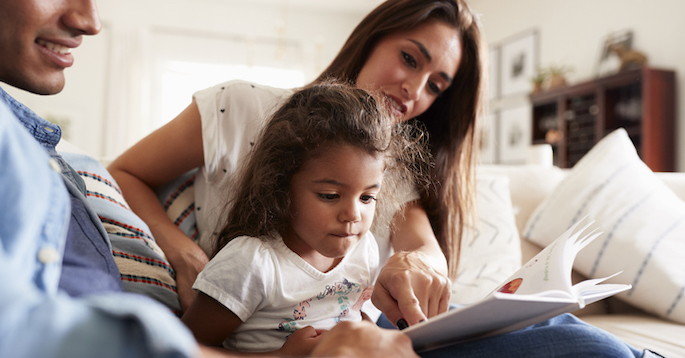 Hispanic couple and their young daughter sitting on the sofa reading a book together at home