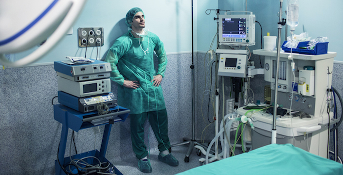 worried or sad looking surgeon leaning against wall of empty operating theater in scrubs