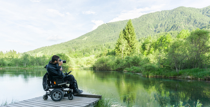 disabled caucasian man using a power wheelchair sitting on a wooden dock taking a photograph of a lake and green trees and hills beyond