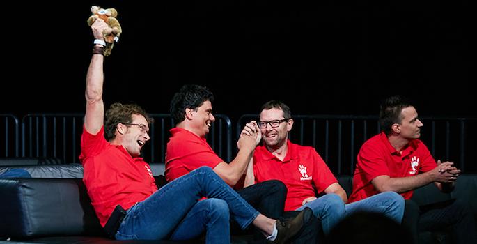 Winners in red team shirts sitting on sofa cheering as results are announced. Volgyesi is triumphantly raising a stuffed toy animal and laughing.