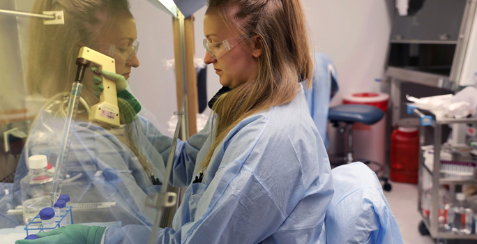 Caucasian woman with blond hair wearing protective clothing runs an experiment housed behind protective plexiglass
