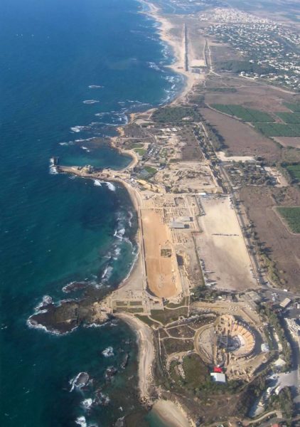 Aerial photograph of ancient site, mostly yellow sand or rock with stone ruins of a semicircular amphitheater, some large swaths of unexcavated land and what may be houses, all abutting the sea