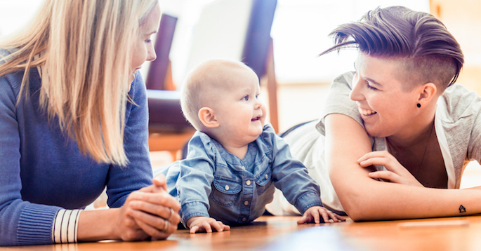 Smiling lesbian couple with baby lying on hardwood floor. Homosexual partners with toddler in living room. Happy family of three at home.