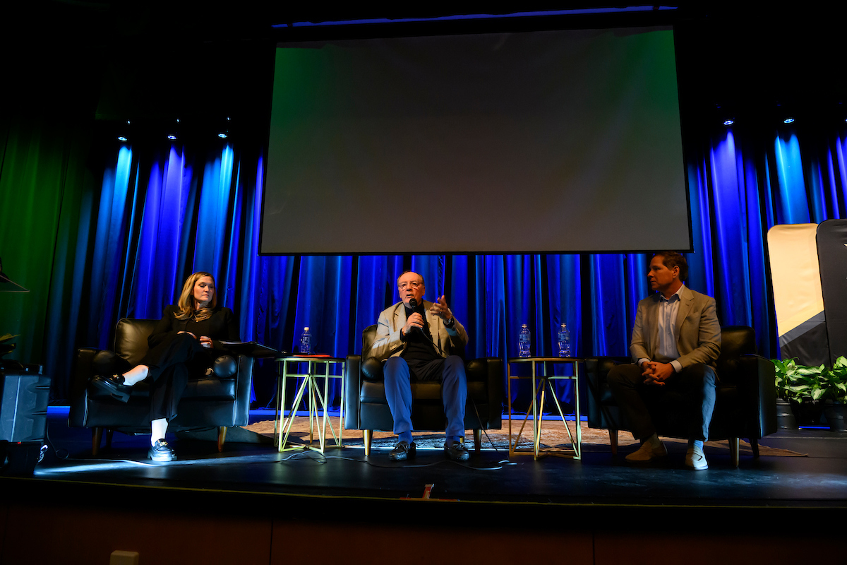 Three speakers seated on a stage against a blue curtain background.
