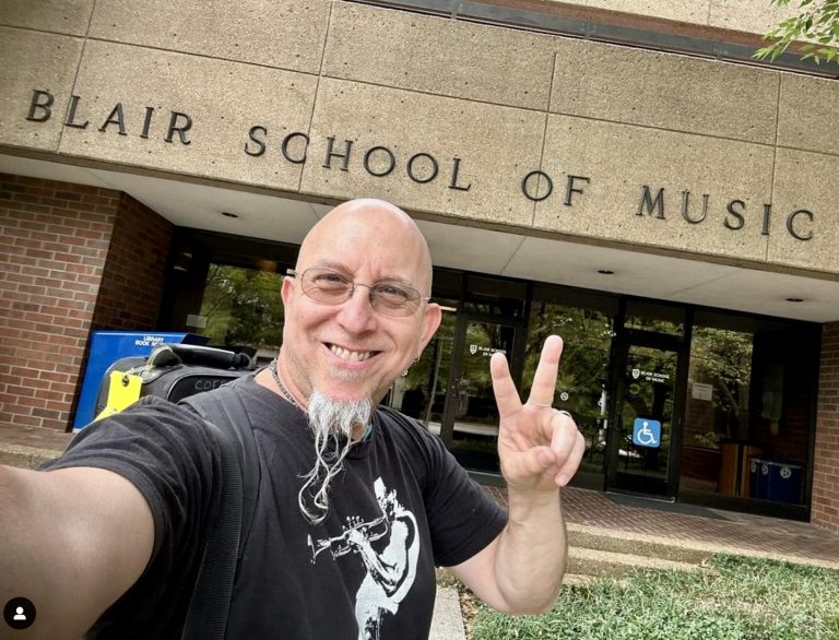 Man wearing glasses and a black t-shirt flashes a peace sign in front of the Blair School of Music building