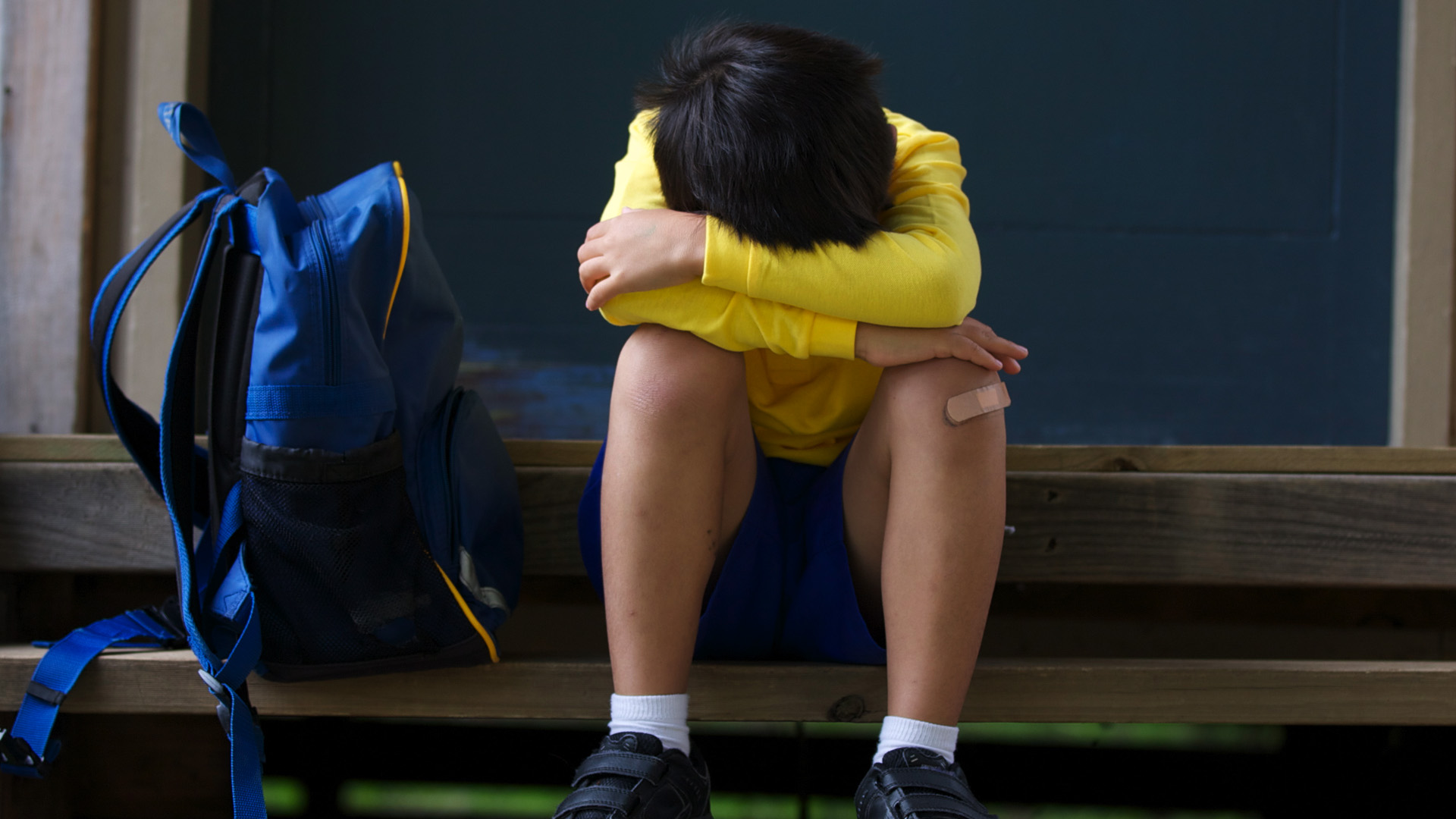 A child sits on a flight of stairs with his head in his arms, appearing to be crying. He has a bandaid on his knee and a blue backpack sits next to him. 