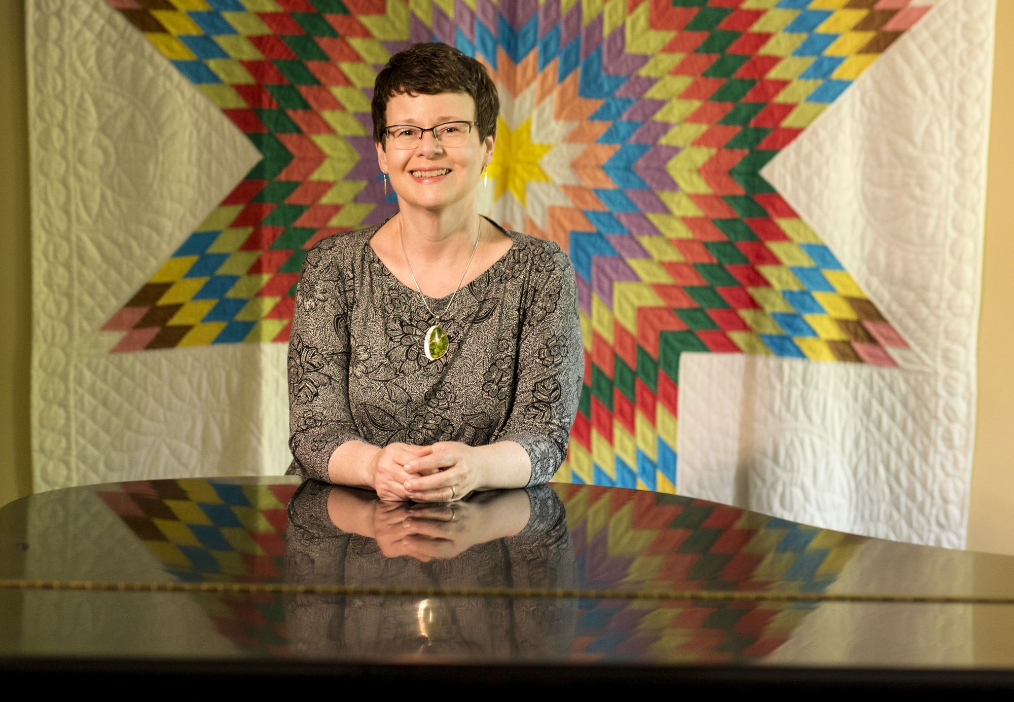 Melissa Rose leans on a piano as she poses in front of a quilted blanket