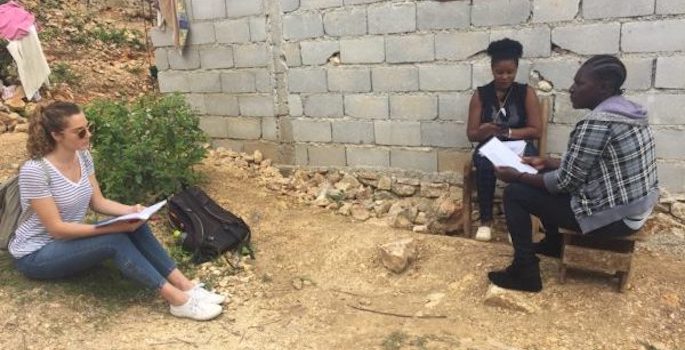 Young caucasian woman sits on the ground facing a black male survey taker and a black woman sitting on a chair outside a cinderblock house