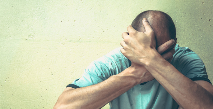 man in 30s, possibly caucasian, sitting against a concrete wall with his head in his hands as if in anguish
