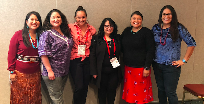 six professional indigenous women, some in cultural dress, smiling in a conference room