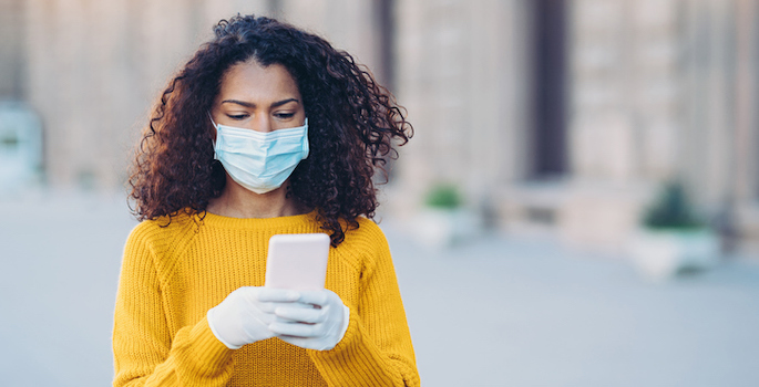 African american woman wearing a face mask walking outdoors and using cell phone