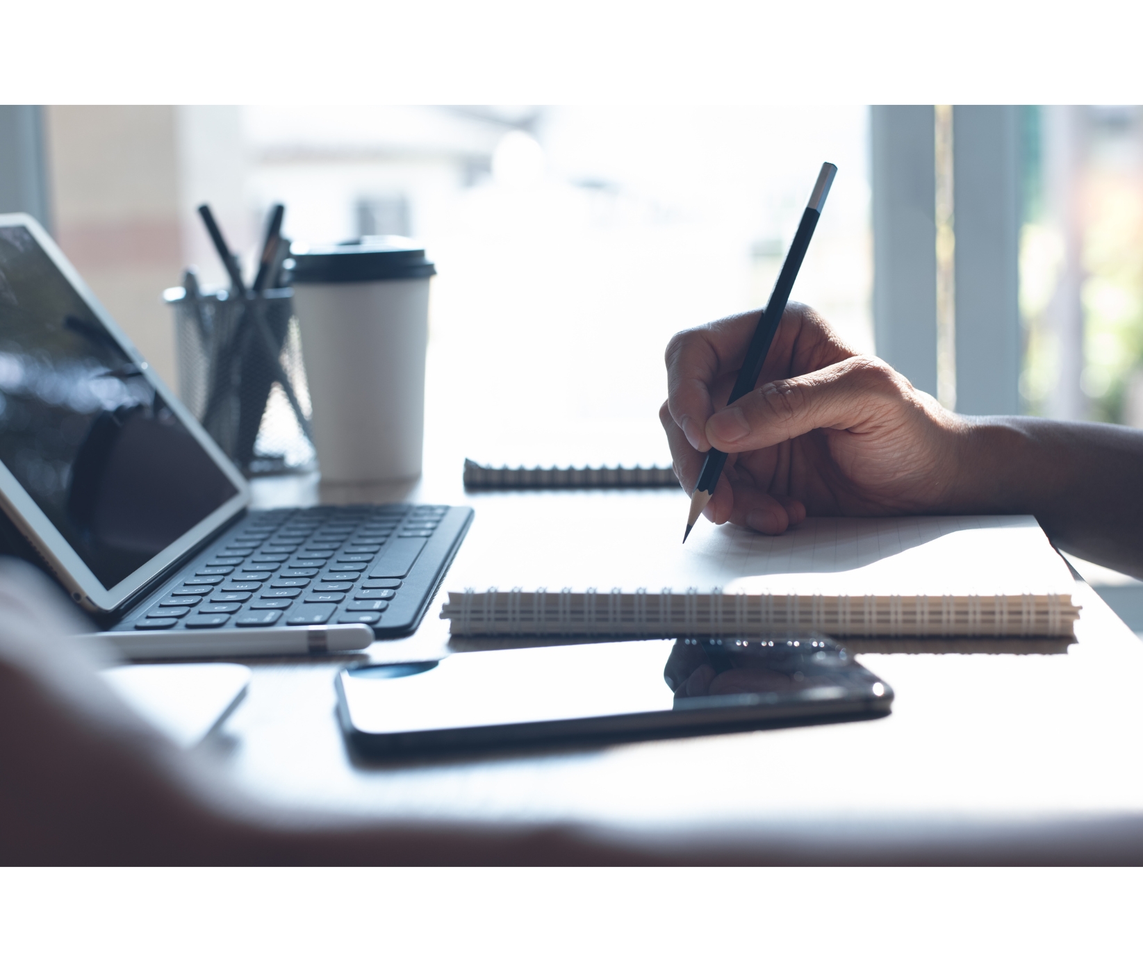 Someone's hand writing with a pencil on paper in front of a laptop computer