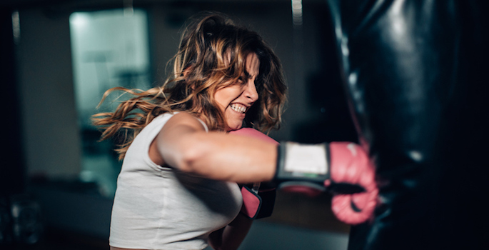 Athletic fair skinned woman with brown hair striking a heavy bag with boxing gloves on