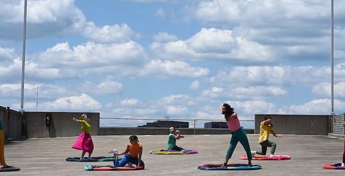 five dancers in colorful street clothes dance within circles delineated by bright soft fabric circles positioned at least six feet apart on the top of a parking garage beneath a bright partly cloudy blue sky
