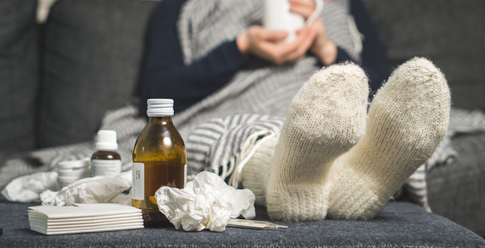 person wearing socks and wrapped in a blanket curled up on sofa with cold medicine