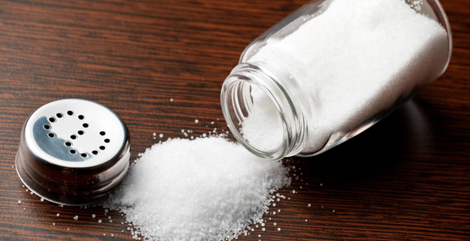 glass salt shaker uncapped and spilled on a brown tabletop