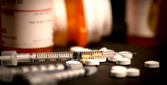 An injectable drug (heroin) is loaded into a syringe while prescription medication is strewn about haphazardly.