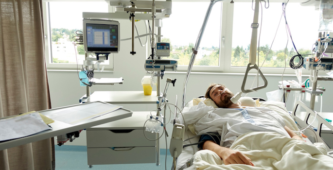 Young man waking up in a hospital bed after surgery. He’s got a breathing tube in his nose and medical equipment around him.