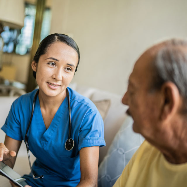 Female nurse sits on a couch with an older male patient having a discussion while using a tablet.