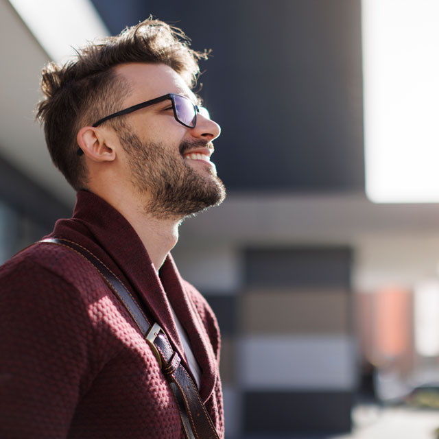 A young man stands outside an urban building with face upturned toward the sky, smiling.