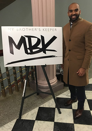 Alumnus Asa Briggs stands next to a sign for My Brother's Keeper