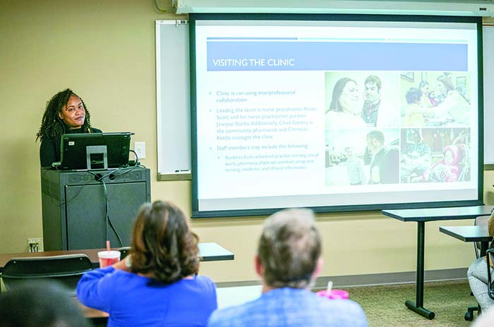 Paris Grady stands behind lecturn next to large screen showing her research results on The Clinic at Mercury Court.