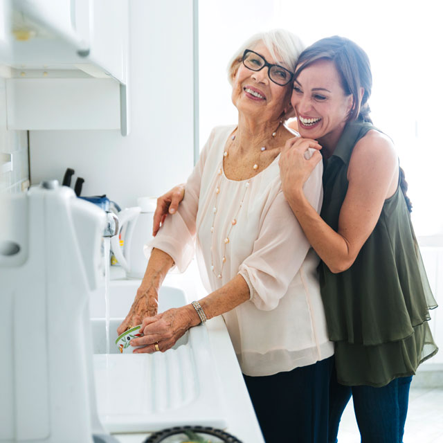 A younger woman hugs an older woman who is washing dishes in a kitchen sink.