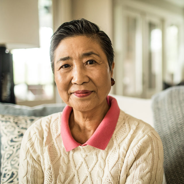 A head and shoulders shot of an older Asian woman sitting on a couch with a big cozy pillow.