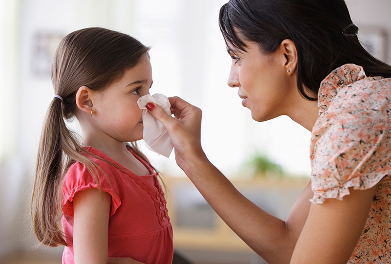 A mother helps her young daughter blow her nose with a tissue.
