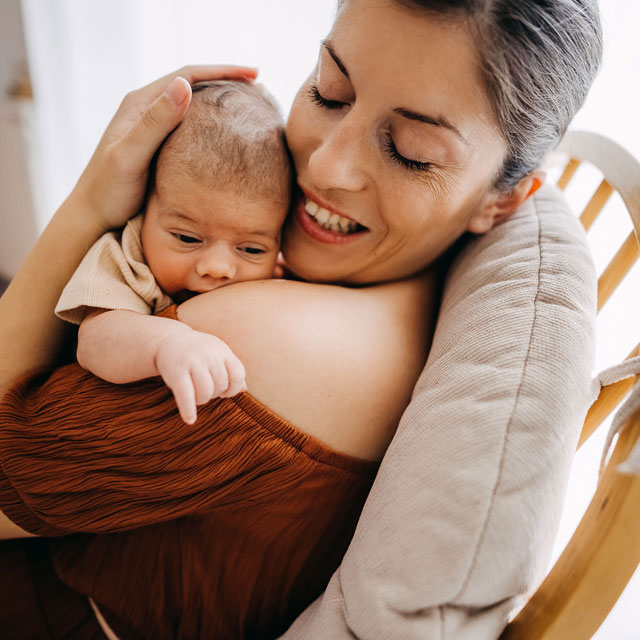 Closeup of a woman in a chair cuddling her newborn on her shoulder.