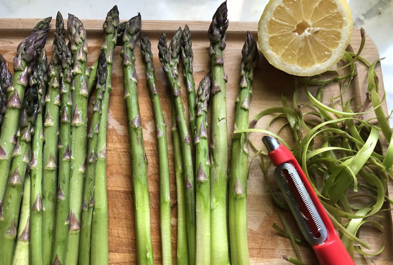 Asparagus spears on a wood cutting board with a peeler and half a lemon.