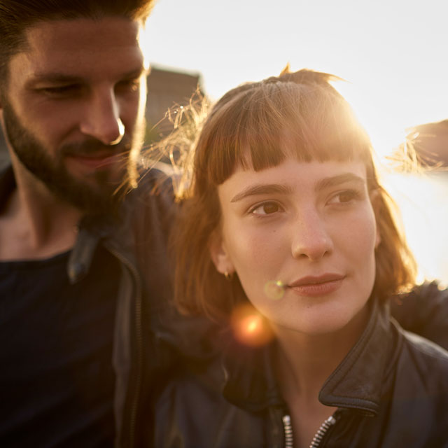 Closeup of a couple, woman standing in front of a man, beside a river in late afternoon light.