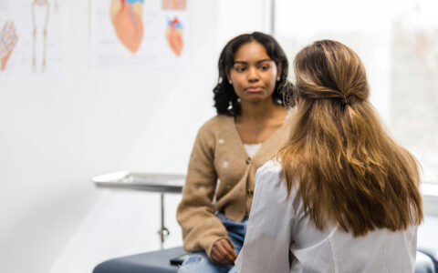 A young woman sits on an examination table at a doctor's office, talking to a female physician.
