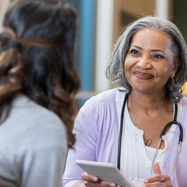 Female doctor or nurse of color faces the camera to talk with a female patient whose back is to the camera.