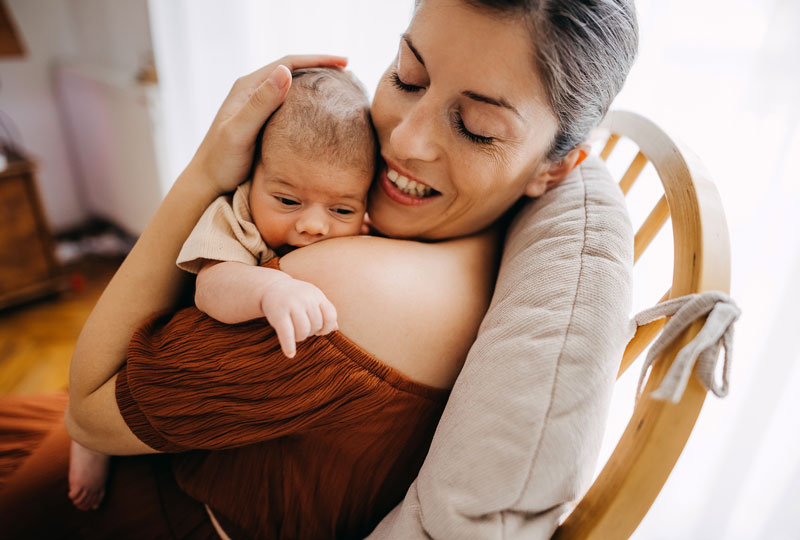 Closeup of a woman in a chair cuddling her newborn on her shoulder.