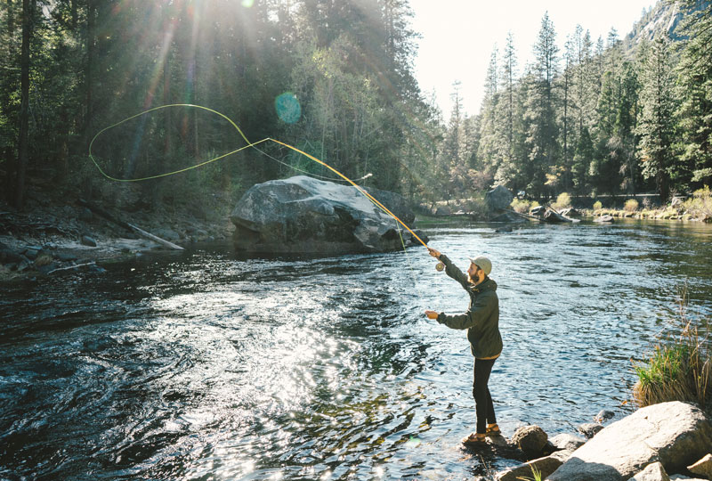 A young man stands at the edge of a river fly-fishing.