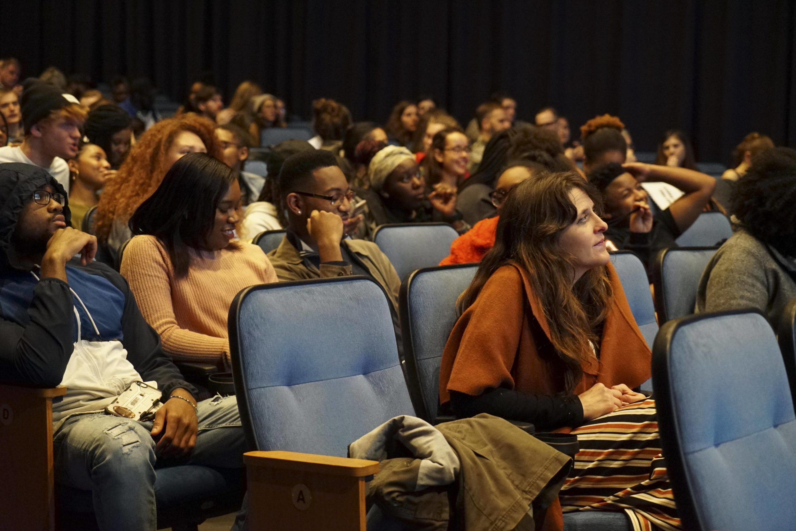A large group of people sit in blue theater seats looking in the same direction
