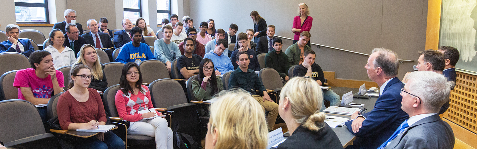 students sitting in chairs listen to a panel of speakers who are sitting at a table at the front of a classroom