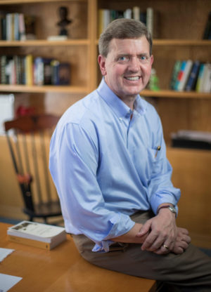 Photo of David Wright sitting on a desk in his office with bookshelves in background