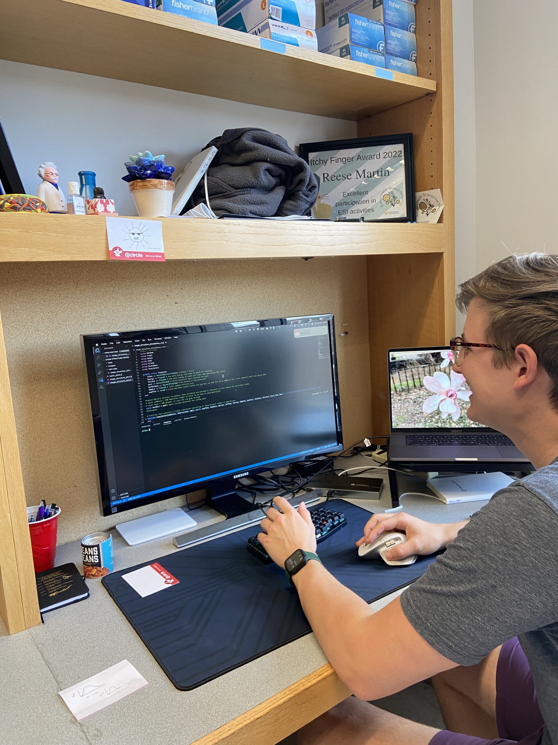 A young man wearing glasses is seated at a desk, working on a computer. He is typing on a keyboard and looking at the code displayed on the monitor. The desk has shelves with various items, including a framed certificate, a potted plant, and some personal belongings. Another laptop is open on the desk, displaying a different screen.
