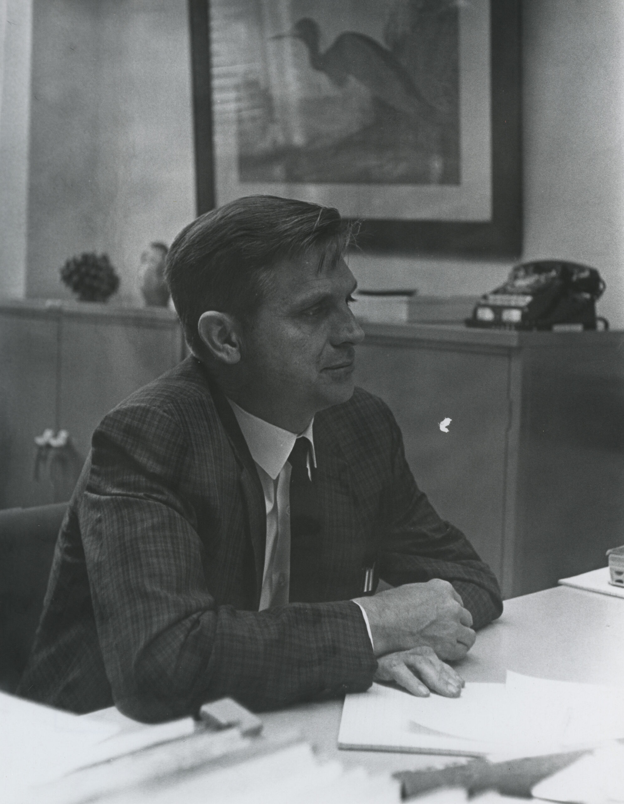 Greyscale image of Ben Channell sitting in his office with a photo of an egret and a rotary phone in the background