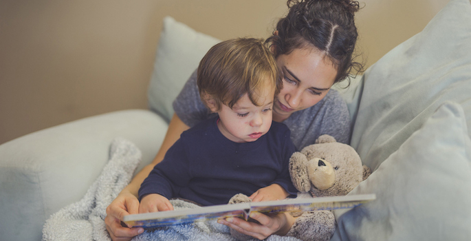 Young Hispanic mom reads a book aloud to her toddler son. They're pointing at the pictures together. A teddy bear is snuggled up with them.