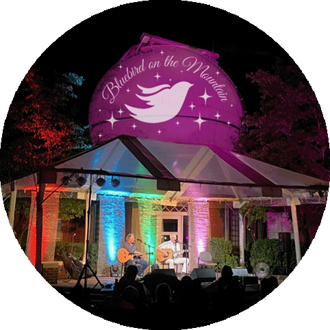 Outdoor concert under colorful lights with musicians on stage, featuring a ''Bluebird on the Mountain'' logo projected above.