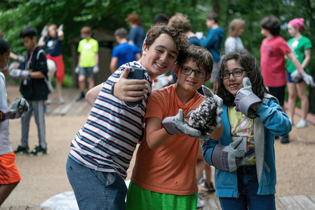 Three smiling kids at camp pose with gloves on, proudly holding a meteorite while others work in the background.