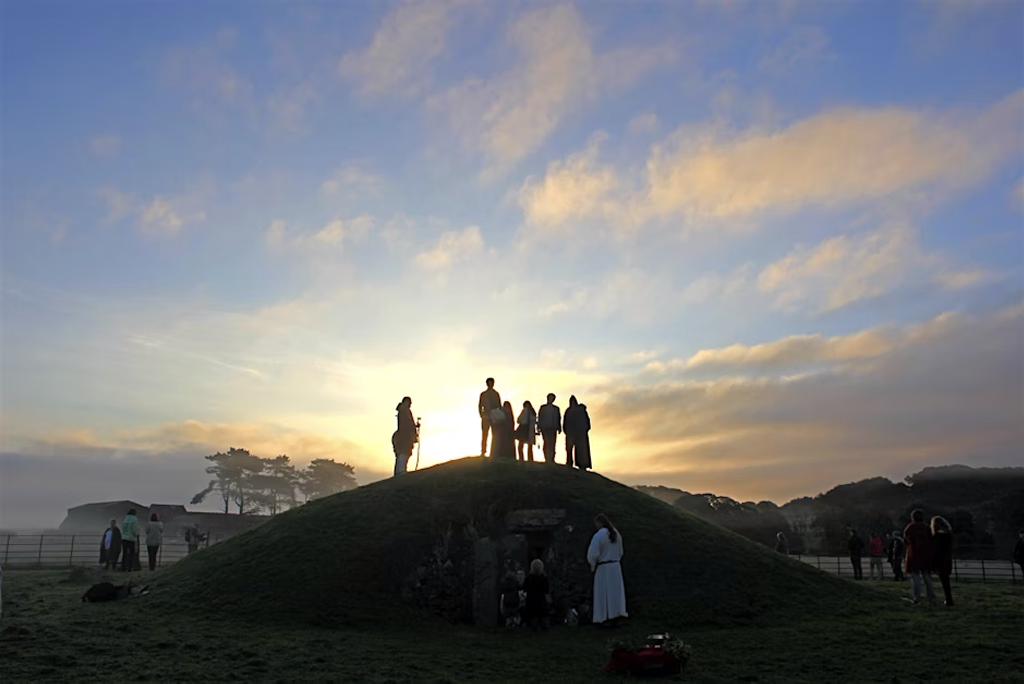 People silhouetted against a sunrise stand atop a grassy mound during a gathering at Bryn Celli Ddu on Summer Solstice.